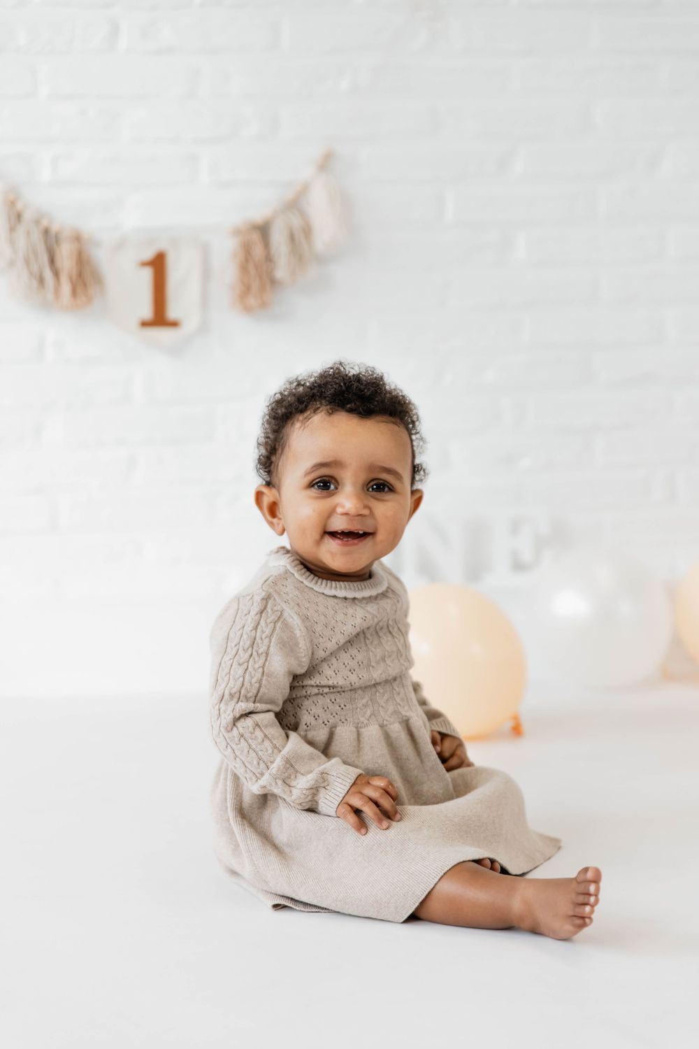Baby in cozy white knit sweater sits on white background with warm lighting and luxurious studio setting.