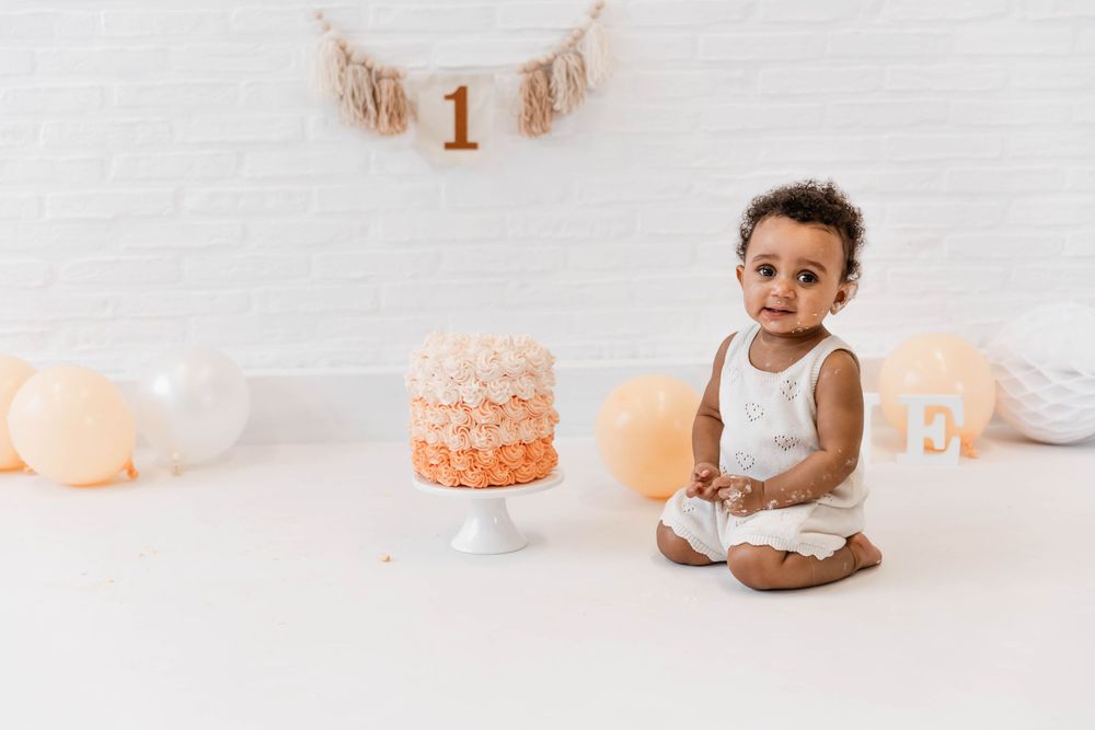 Simple white backdrop first birthday setup with an ombre peach cake on a stand and glowing lanterns around.