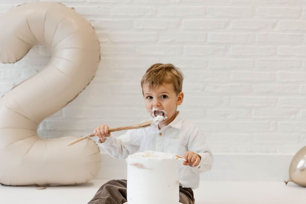 Child sits on floor with balloon number 2 in background while eating from white bowl with spoon.