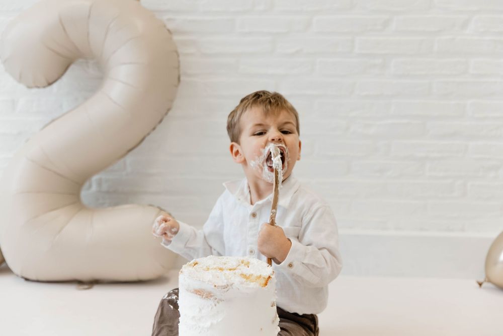A small child in white smiles while playfully eating birthday cake with frosting on their face.
