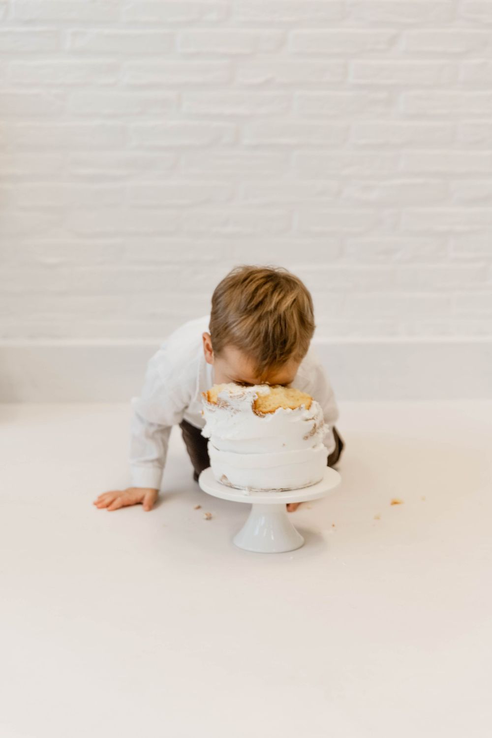 A toddler wearing gray leans over a white cake on a pedestal stand against a plain white background.