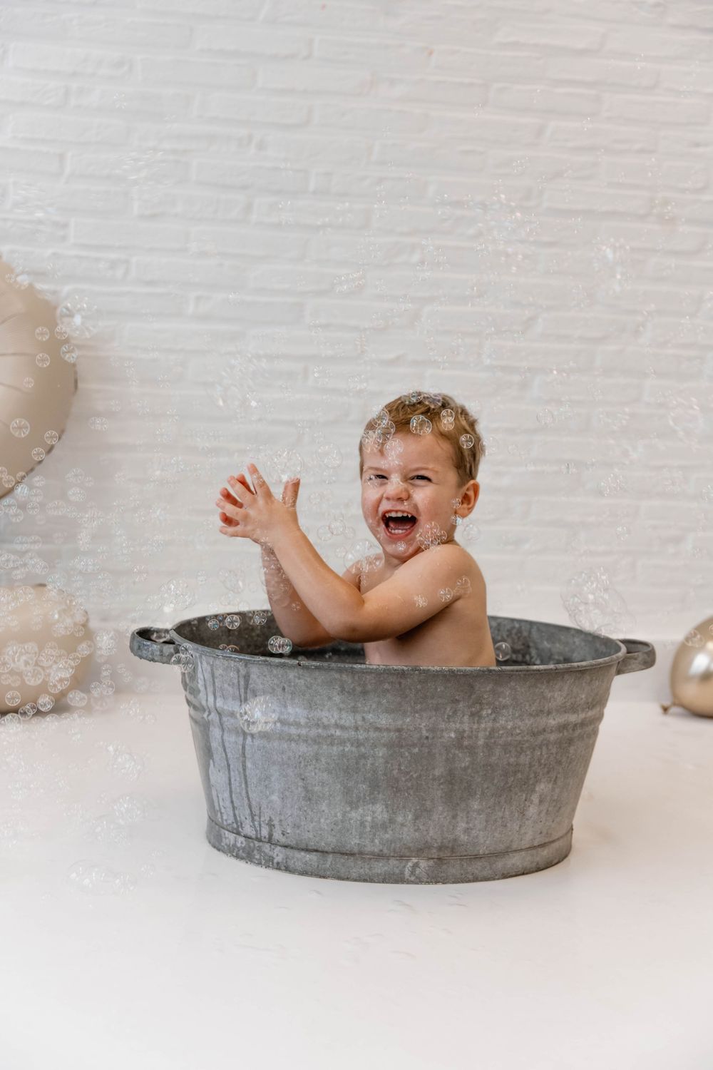 Small child laughs joyfully while taking bath in vintage metal washtub against white brick wall with balloons.