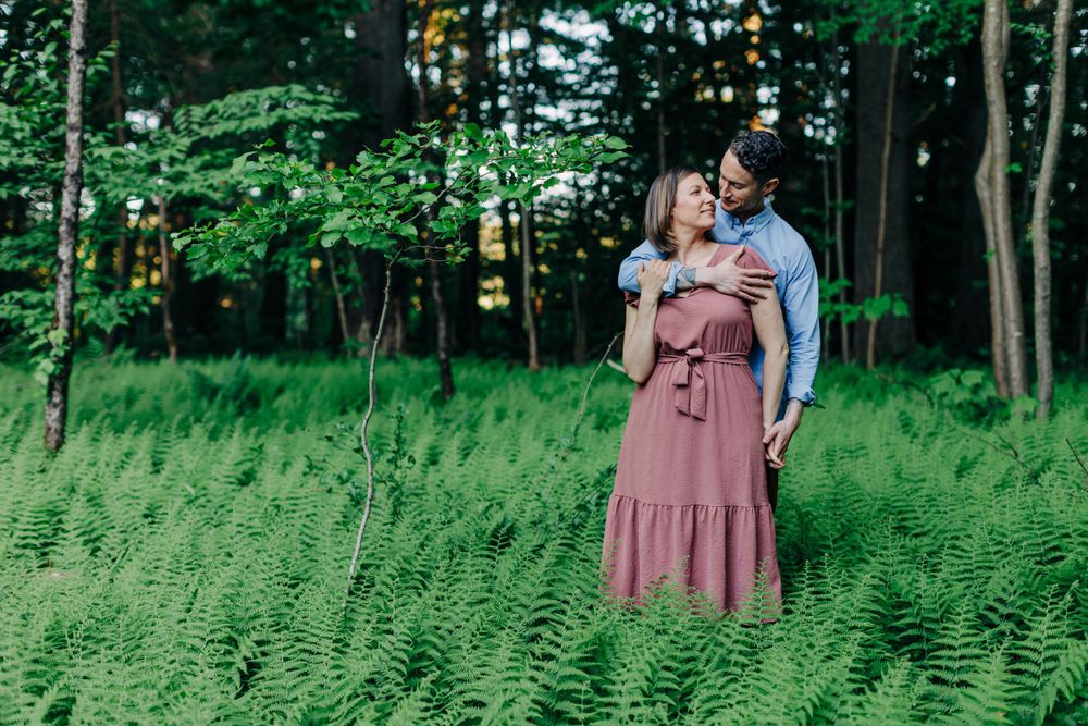 A couple shares an intimate moment while standing in a lush green field surrounded by forest trees.