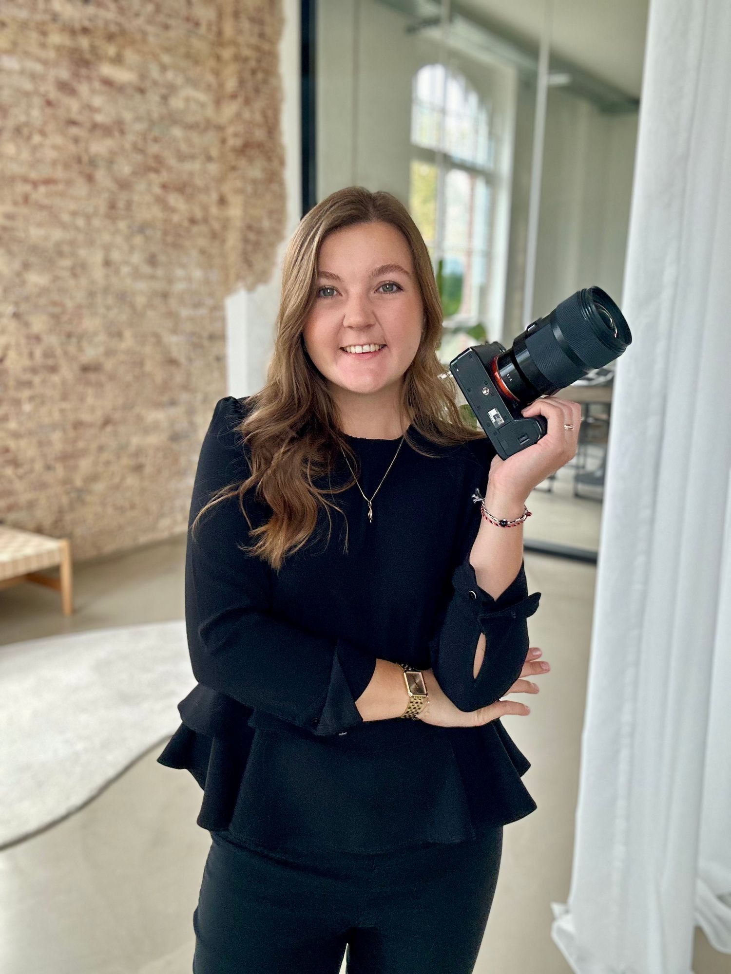 A photographer in a black sweater poses playfully with a camera in a bright studio space with exposed brick walls.