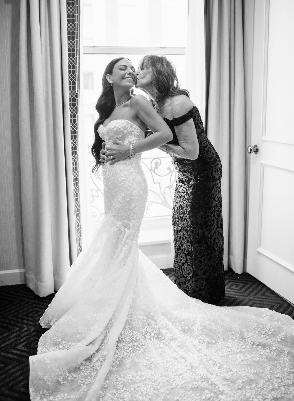A candid black and white wedding moment showing a strapless lace gown and black formal dress during preparation.