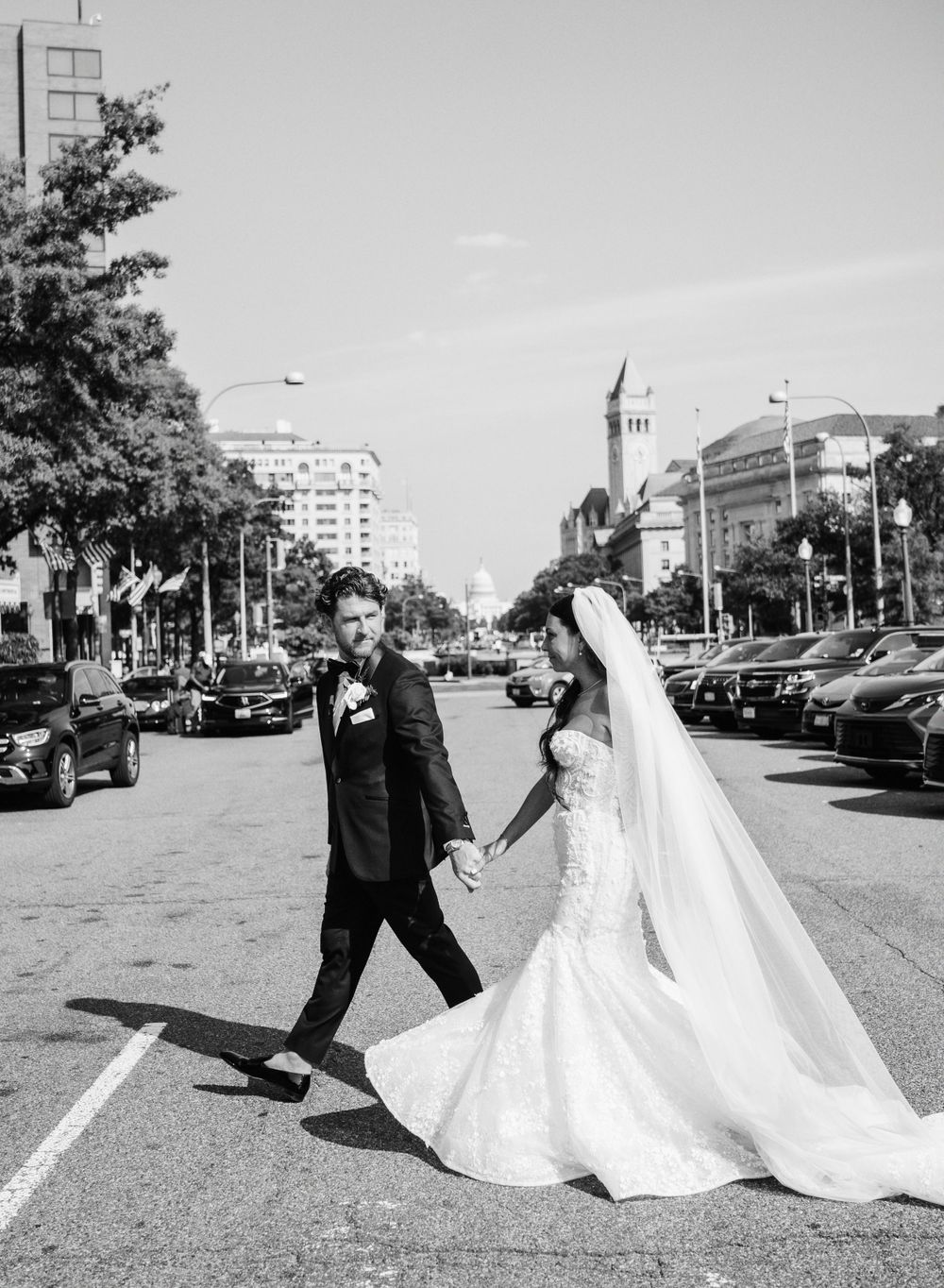 A black and white photo of newlyweds walking down a city street with classic architecture in the background.