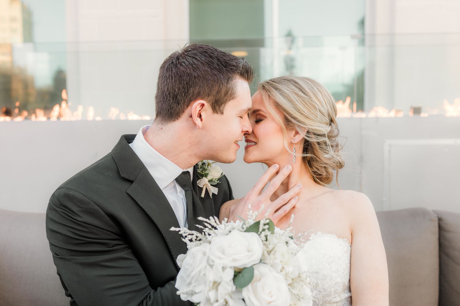 A romantic wedding moment with a bride in white holding a floral bouquet and groom in black suit sharing a tender kiss.