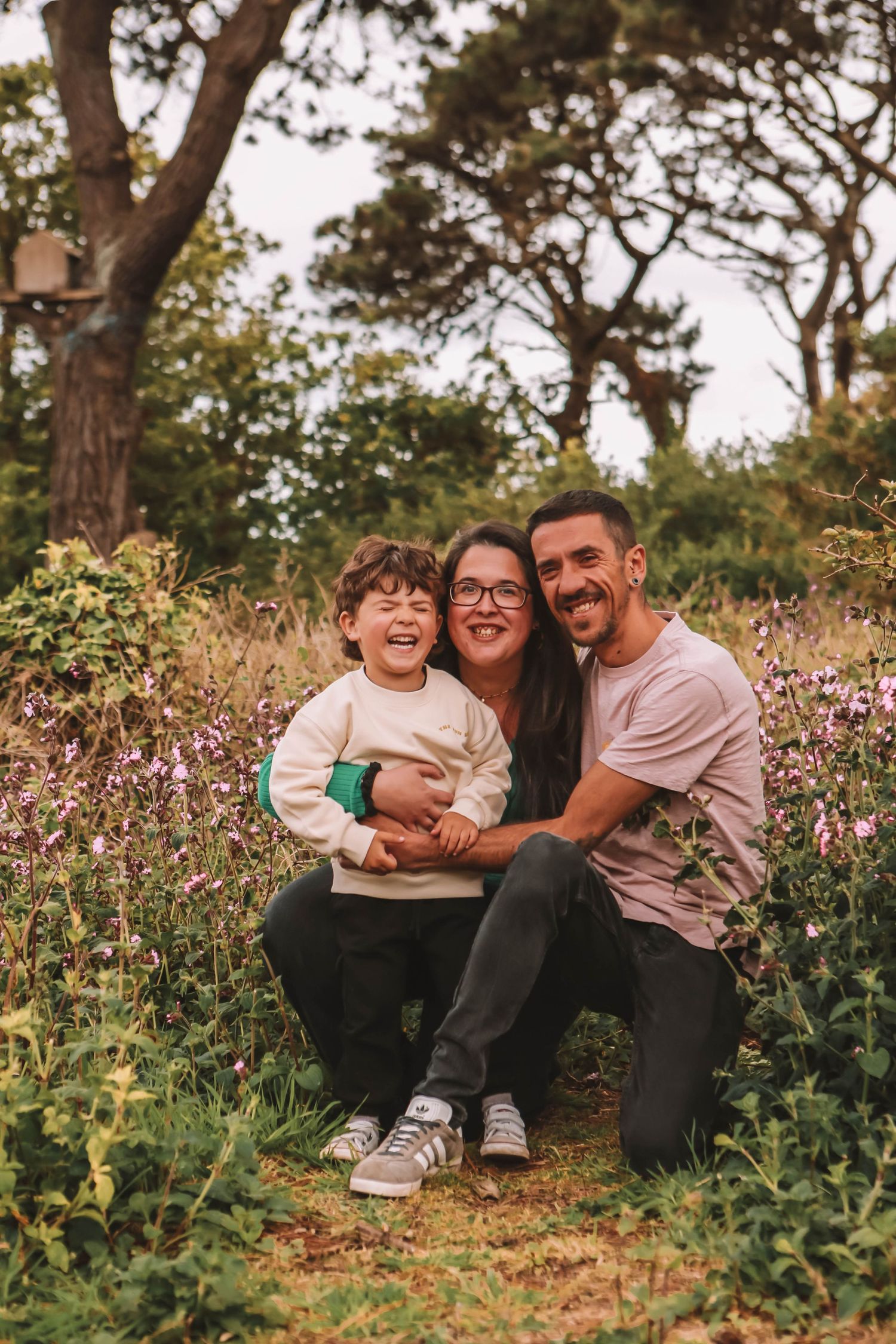 Happy family of three posing outdoors among wildflowers and trees, smiling and laughing together.
