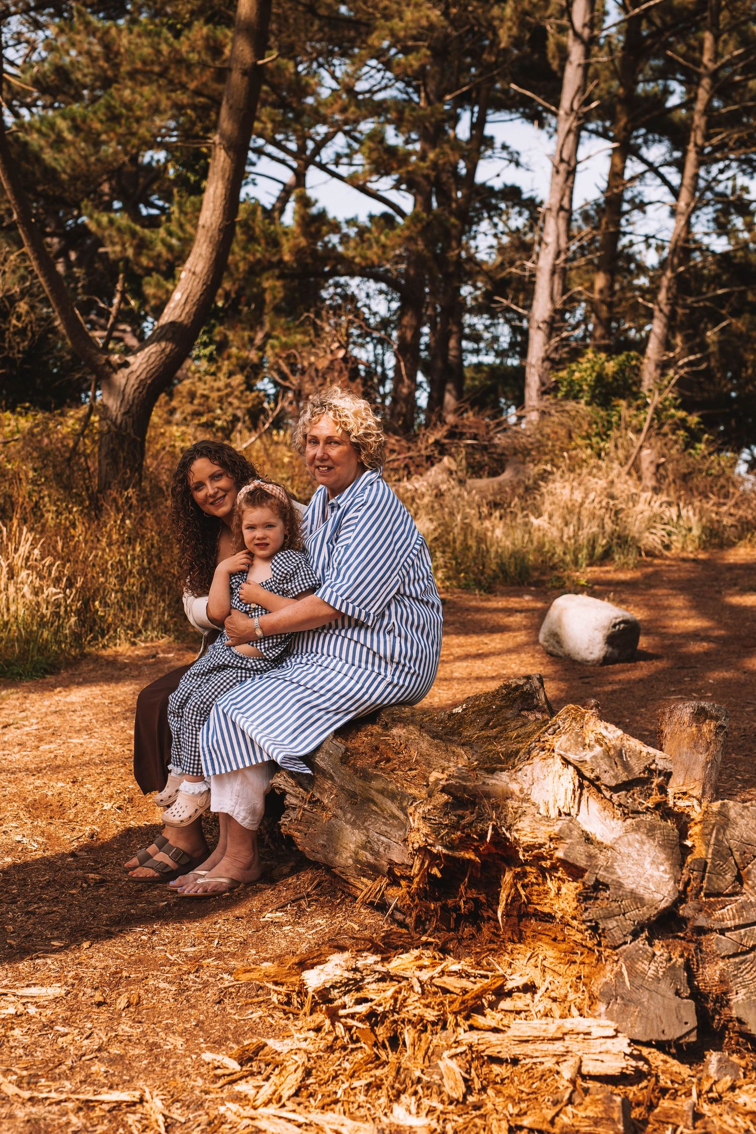 A parent and child sit together on a fallen log in a sunlit forest during golden hour.