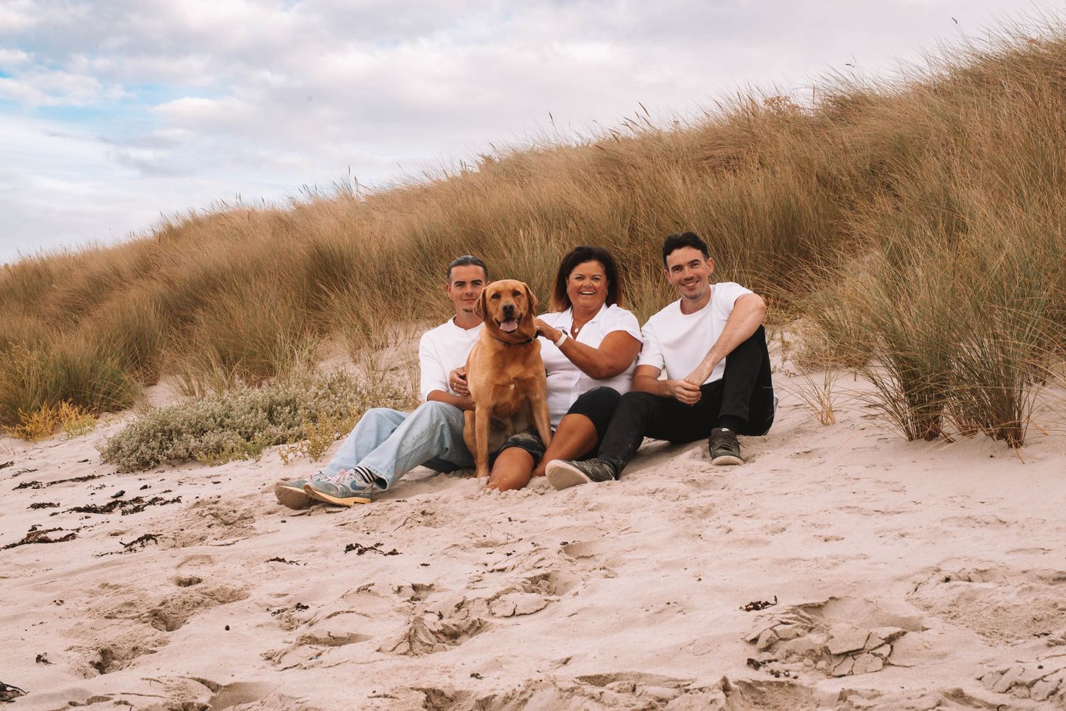 Family of three with a golden dog sitting on sandy beach dunes with tall grass under a cloudy sky.