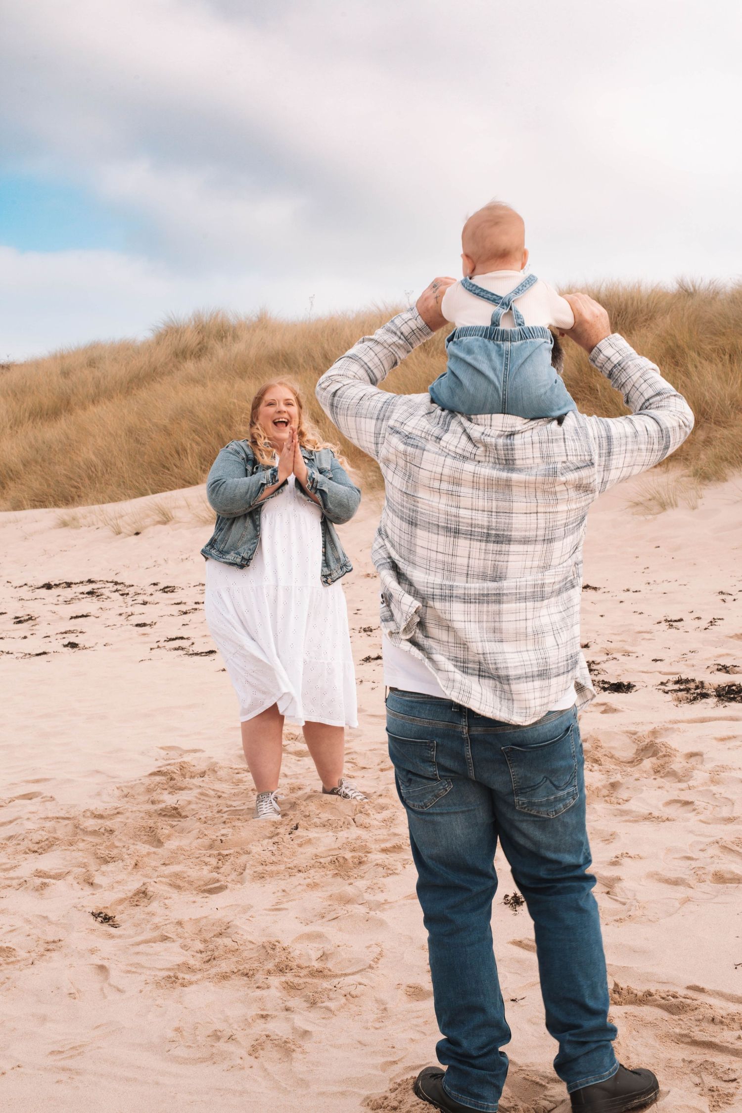 Two people share a playful moment on a sandy beach with coastal dunes in the background.