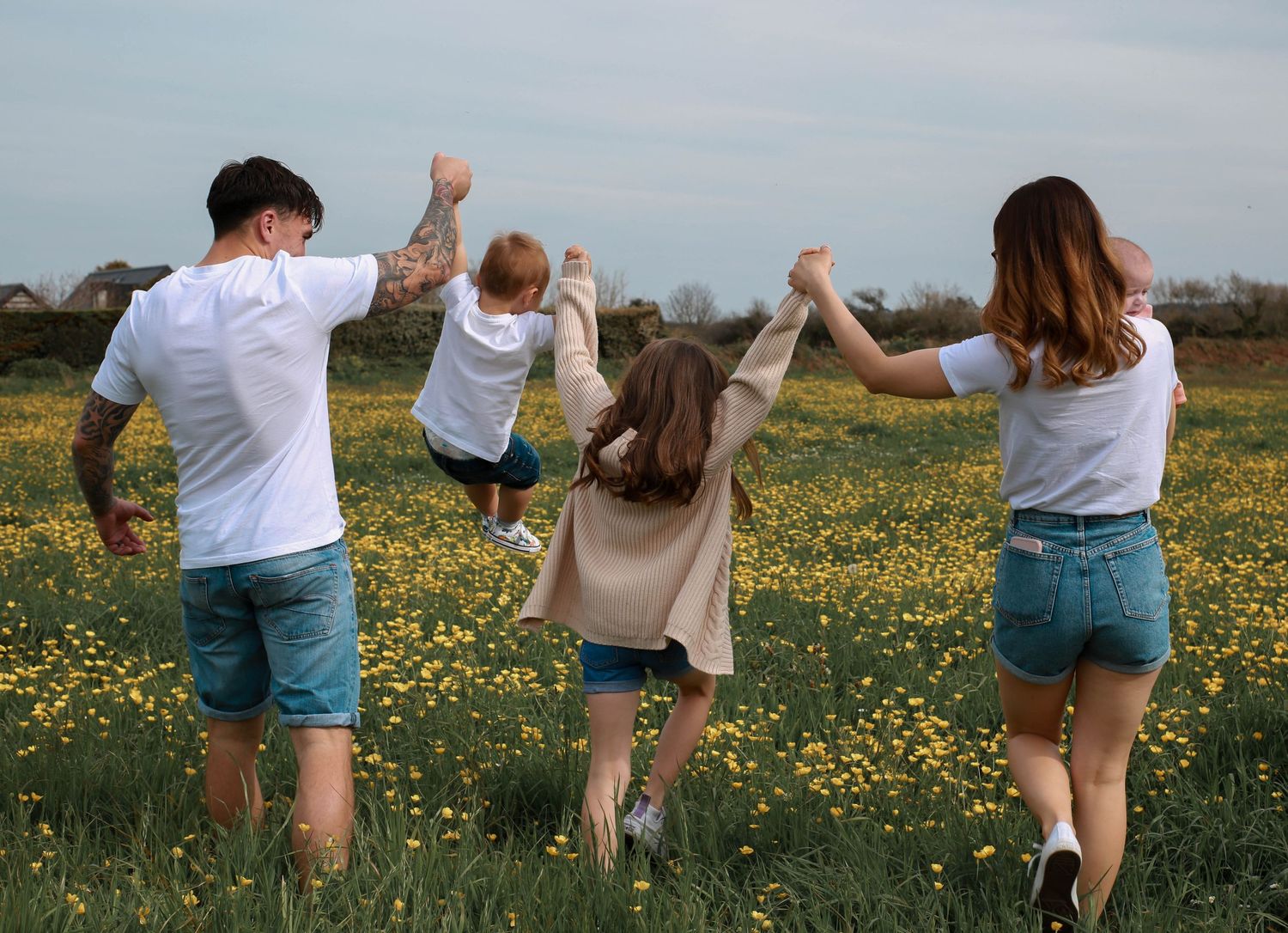 Family of four walking through a sunny wildflower meadow holding hands, viewed from behind on a spring day.