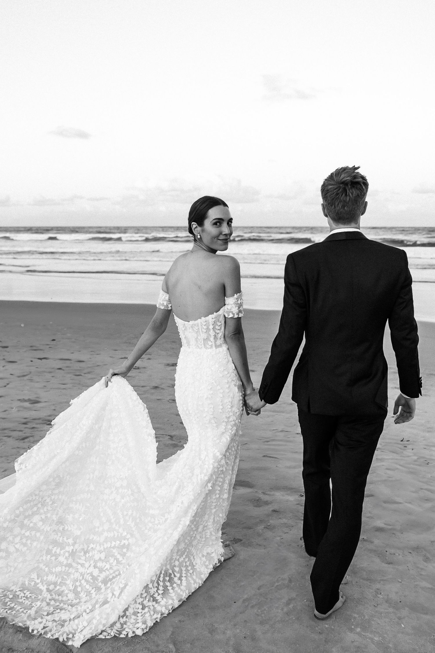 Newlyweds walking hand in hand on a beach, with bride's lace dress trailing in the sand.
