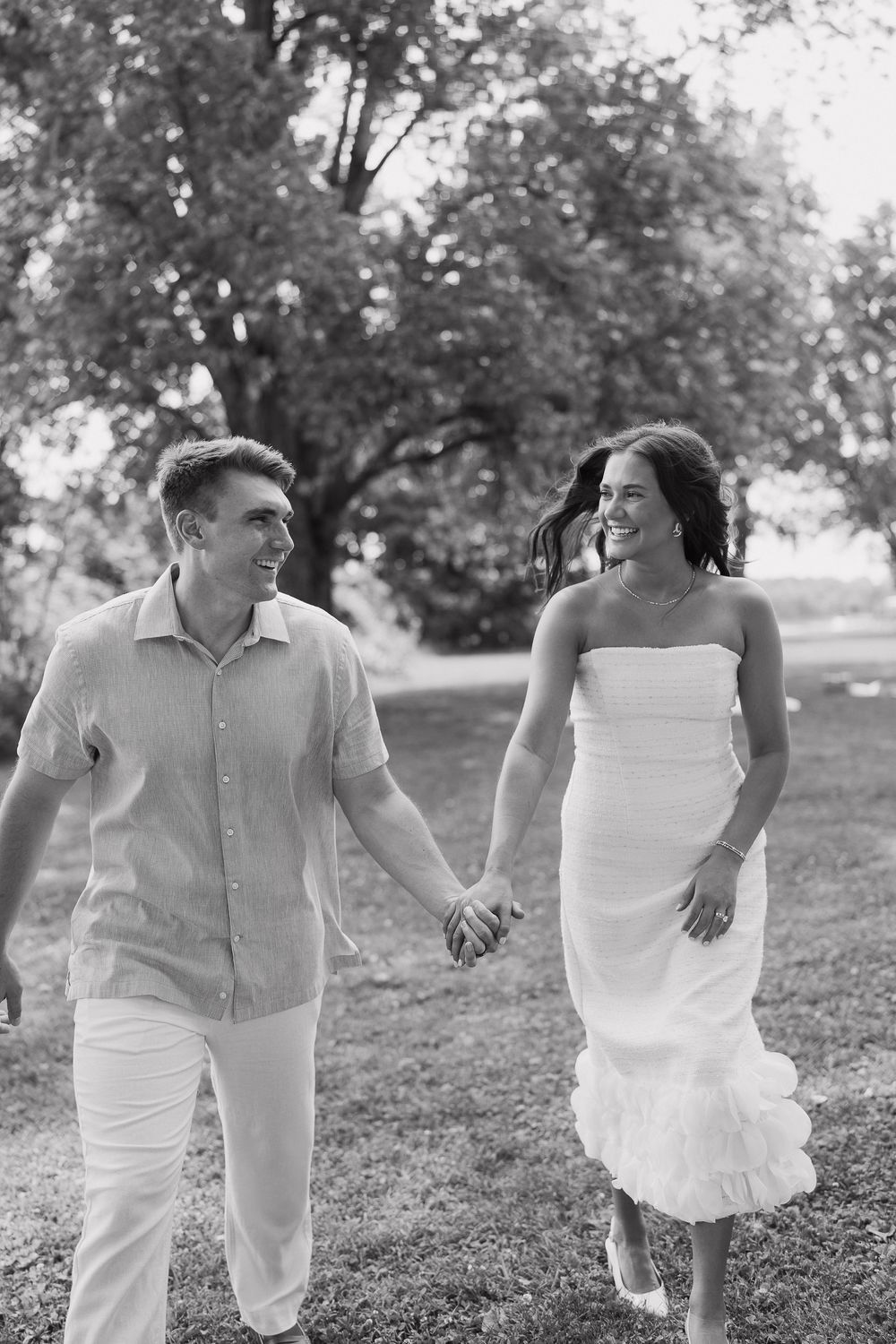 Black and white photo of smiling couple holding hands and walking together through lush green park.