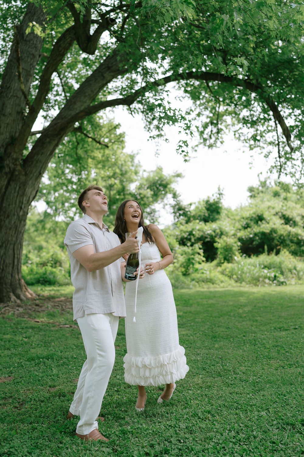Joyful couple in white outfits celebrating outdoors, popping champagne under a large green tree.