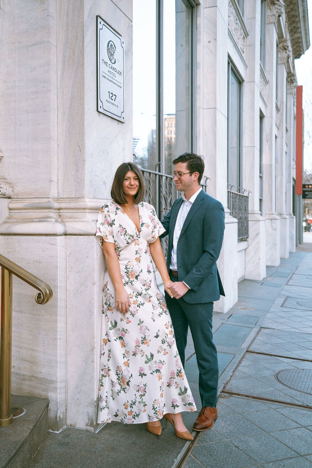 Couple holding hands beside elegant building exterior, woman in floral maxi dress, man in navy suit.