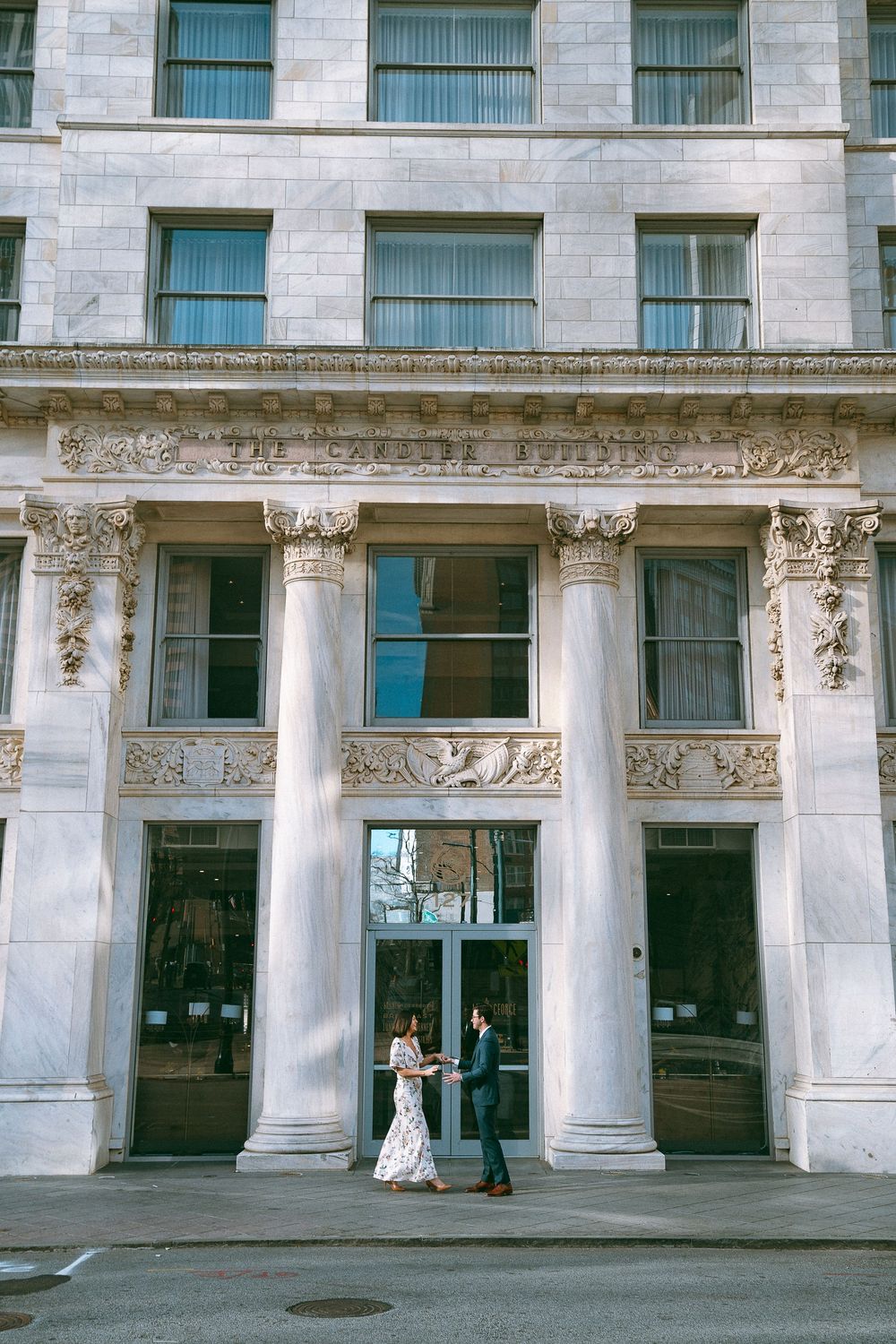 Couple holding hands in front of a grand neoclassical building with ornate columns and decorative frieze.