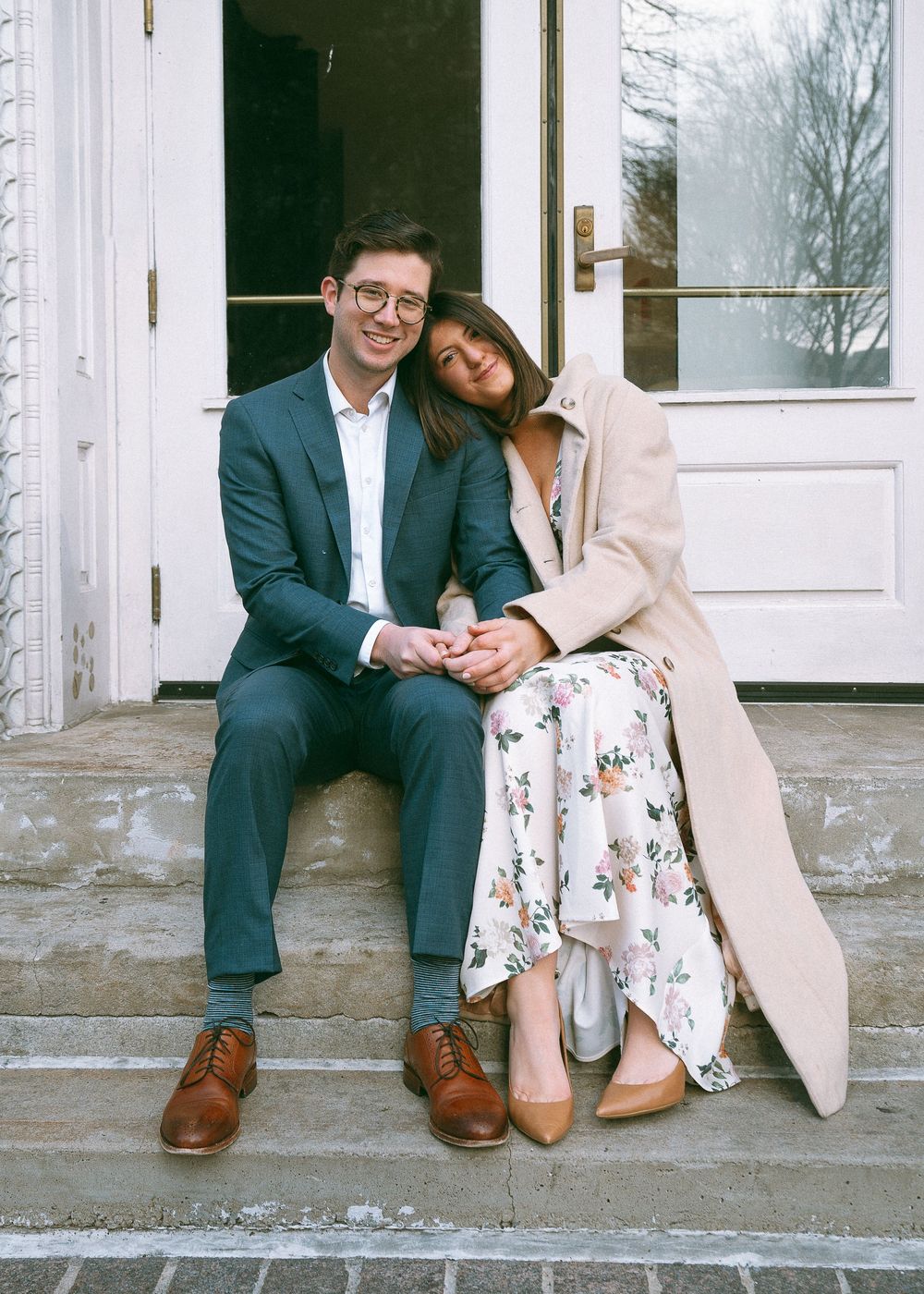 Smiling couple sitting on stone steps, woman in floral dress and beige coat leaning on mans shoulder.