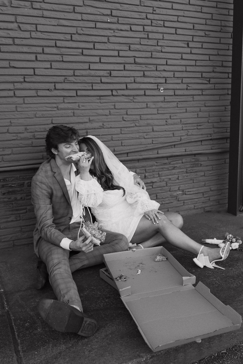 Black and white photo of bride and groom sitting against brick wall eating pizza after wedding.
