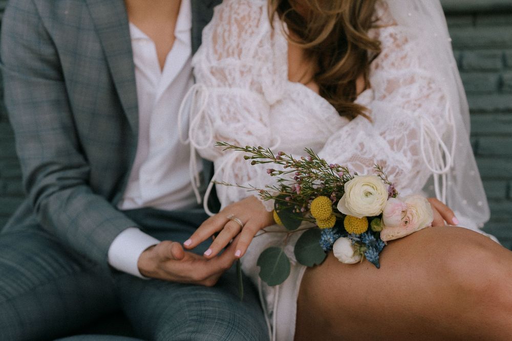 Close-up of bride in lace dress holding a wildflower bouquet while sitting with groom.