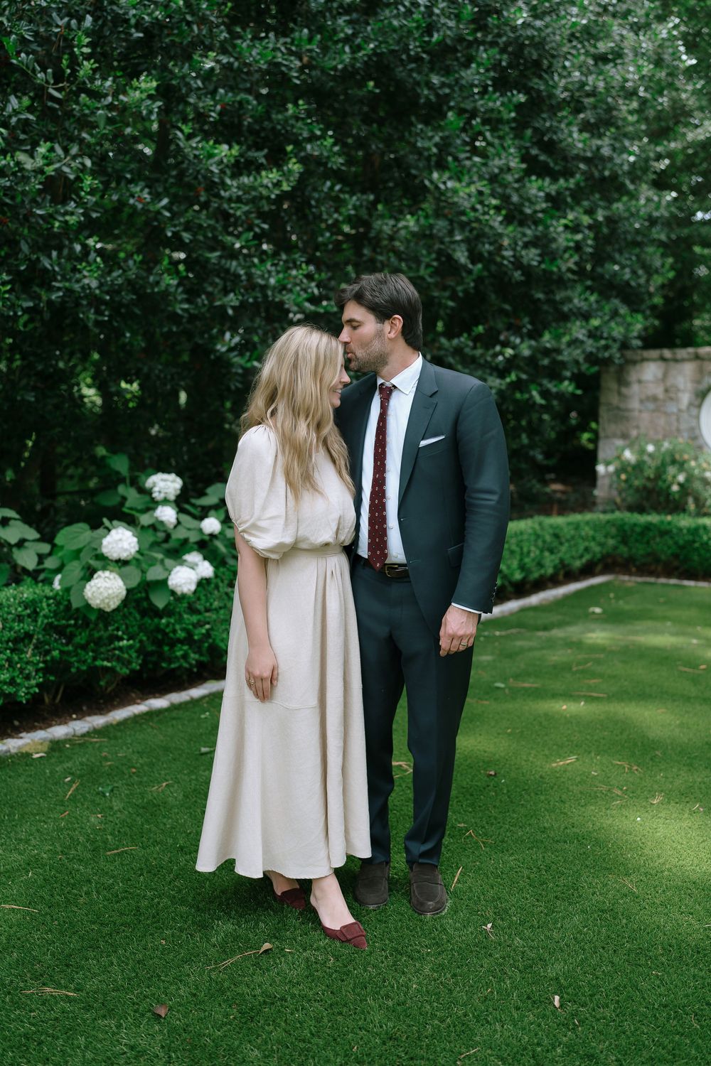 A couple shares a tender moment in a lush green garden, man in dark suit with floral tie, woman in cream dress.