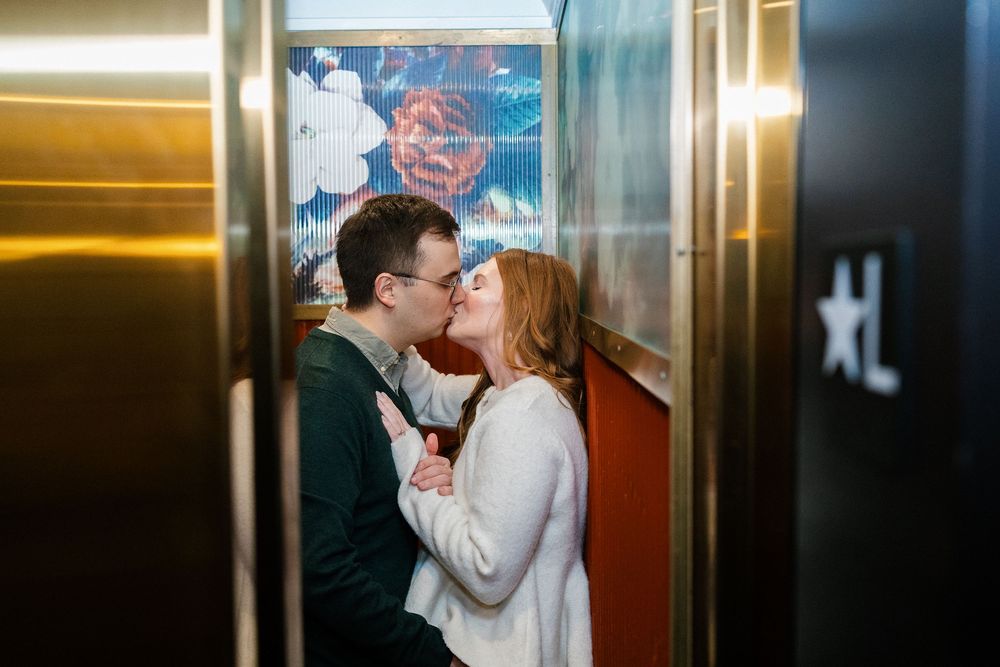 Couple sharing a romantic kiss inside a hotel elevator with colorful artwork backdrop.