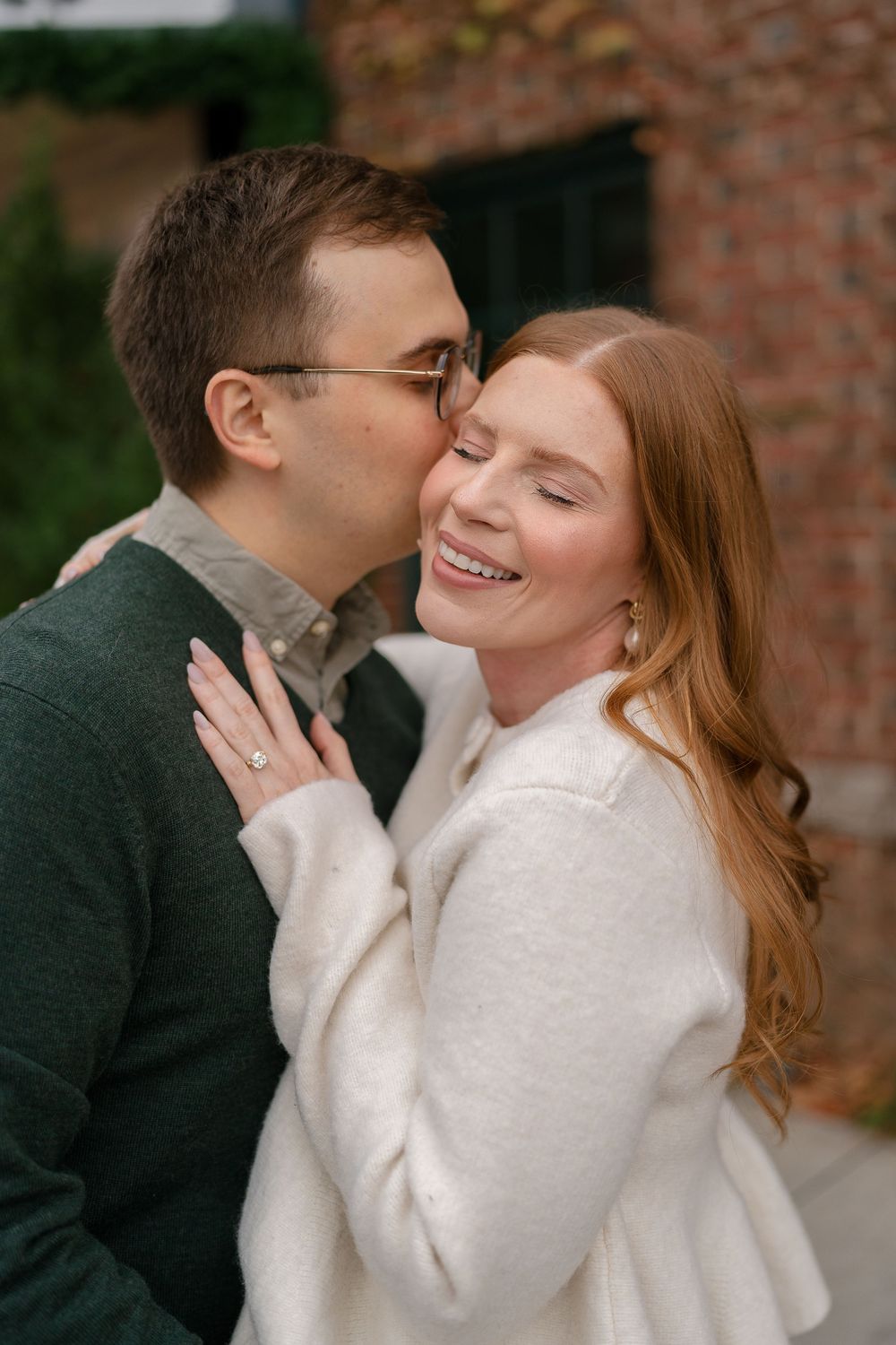 Man in green sweater kissing smiling redhead woman wearing engagement ring outdoors.