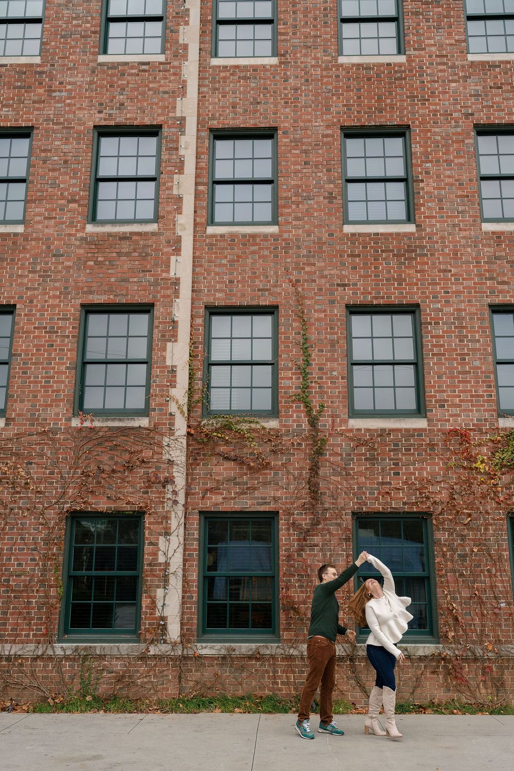 Joyful couple dancing and twirling in front of large brick building with ivy.