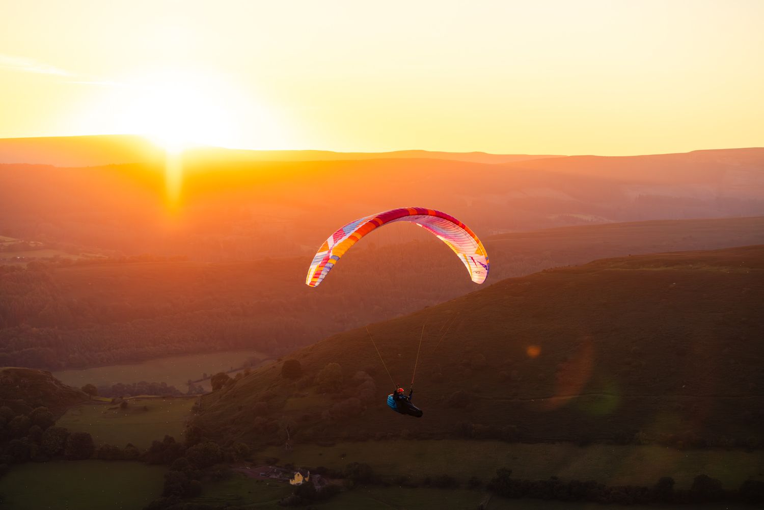 Paraglider soaring against vibrant orange sunset over rolling hills and valleys.