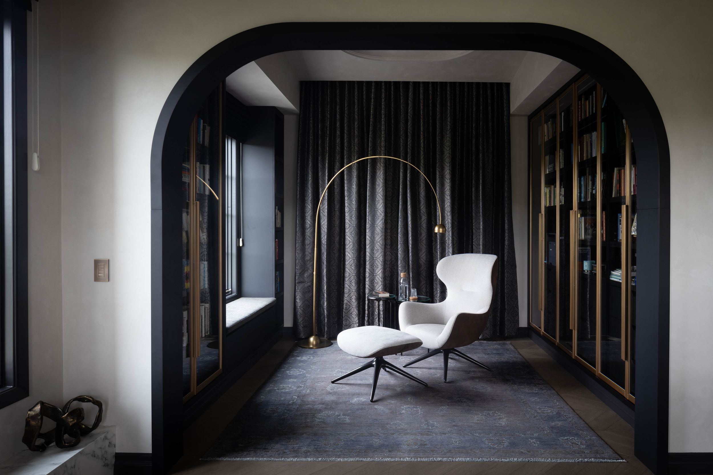 Elegant dark interior with arched black doorway, white lounge chair, ottoman, gold arc lamp, and floor-to-ceiling curtains.