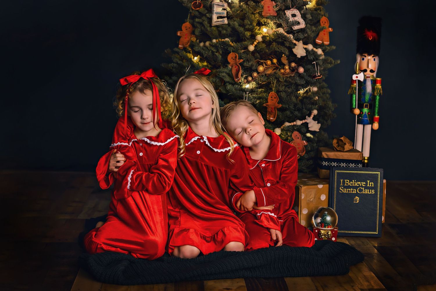 Three children in matching red holiday sleepwear pose in front of a decorated Christmas tree.