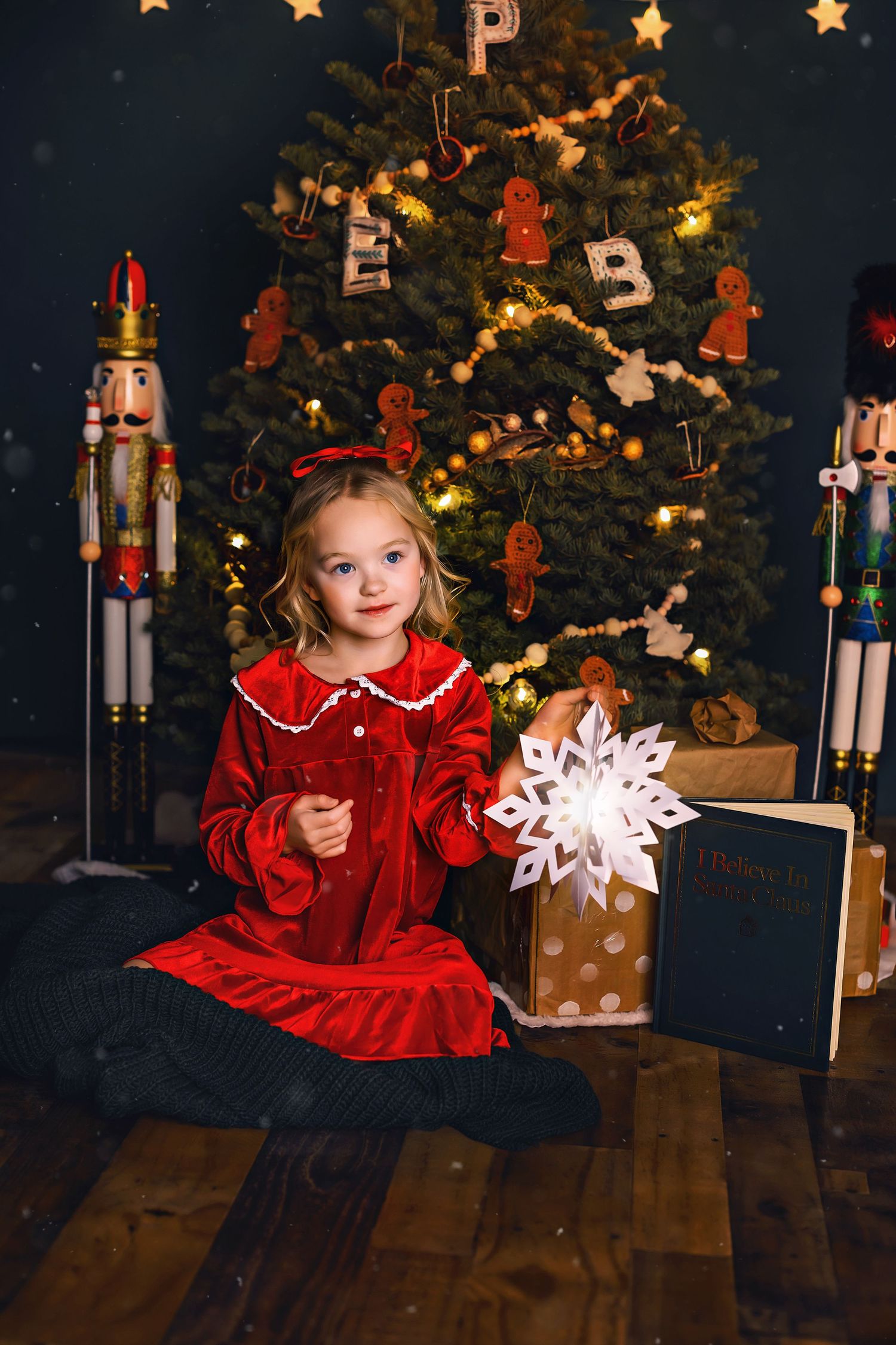 Child in red velvet holiday dress holds a snowflake decoration by a Christmas tree with nutcrackers.