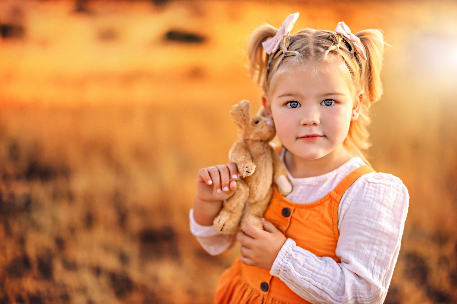 A playful child in orange overalls holds a teddy bear against a warm autumn background.