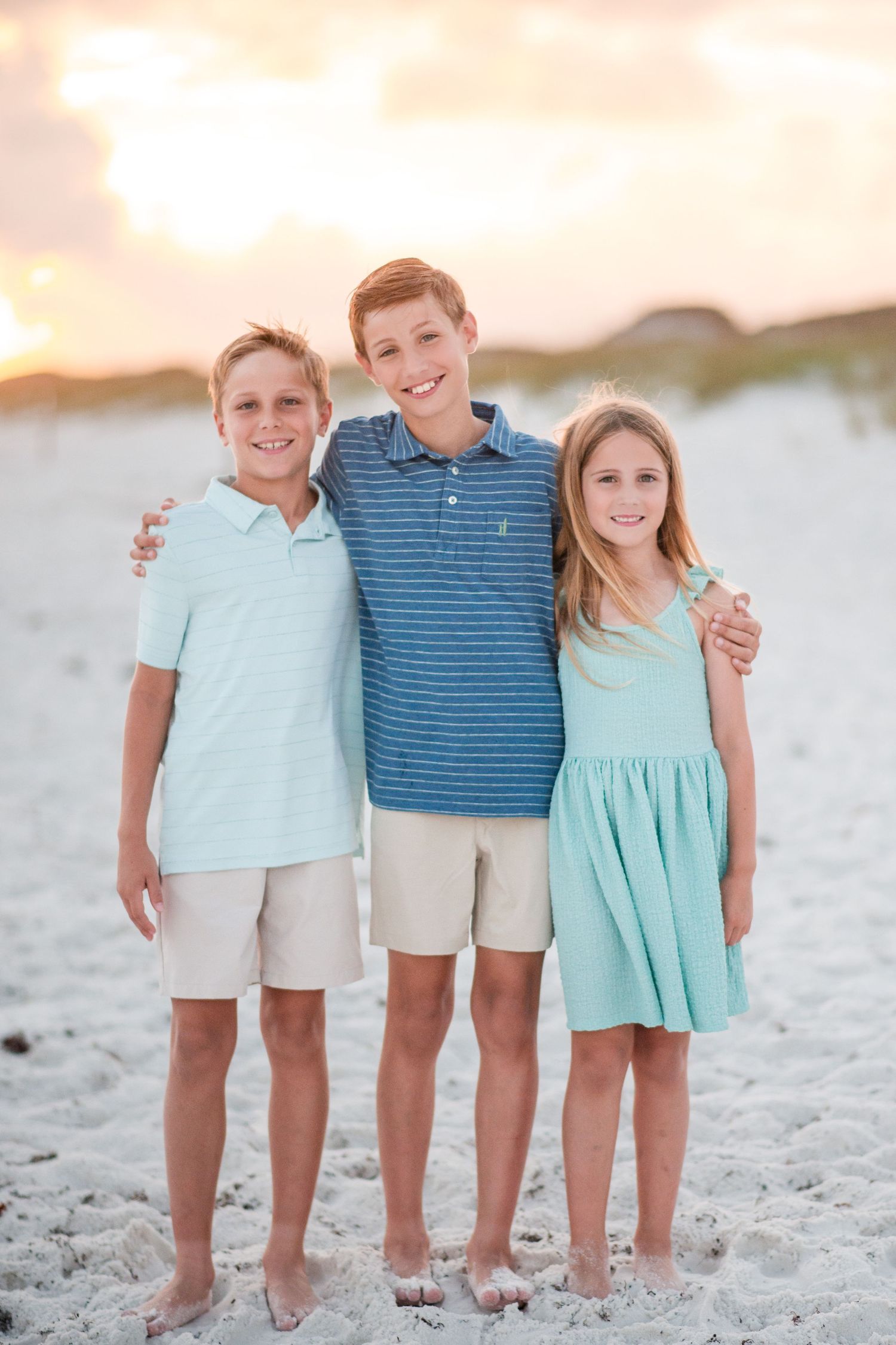 A family of five poses together on a beach at sunset wearing coordinated blue and coral outfits.