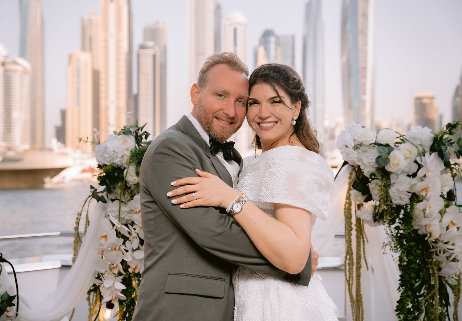 A wedding couple embraces on a balcony overlooking a city skyline with white floral decorations.