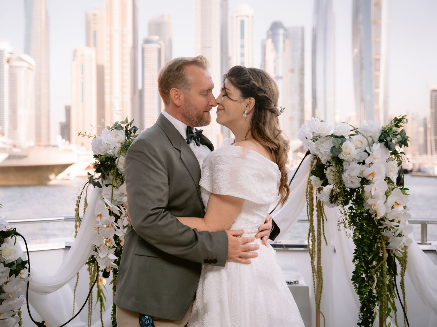 A romantic wedding couple embraces on a rooftop terrace decorated with floral garlands and the city skyline in the background.