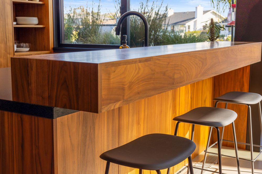 Modern kitchen island with sleek wooden paneling and black bar stools features a dark countertop and window view.