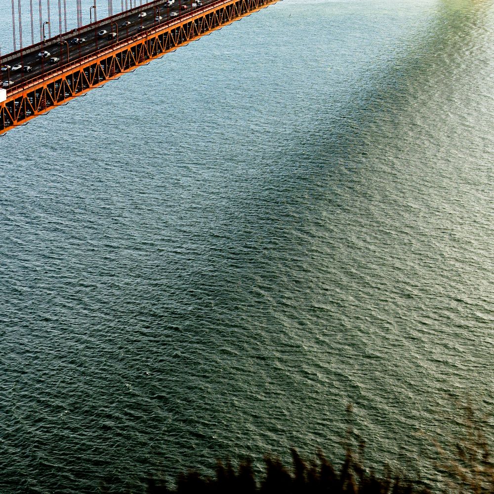 Aerial view of a bridge structure spanning across rippling green-blue water with slight sunlight reflection.