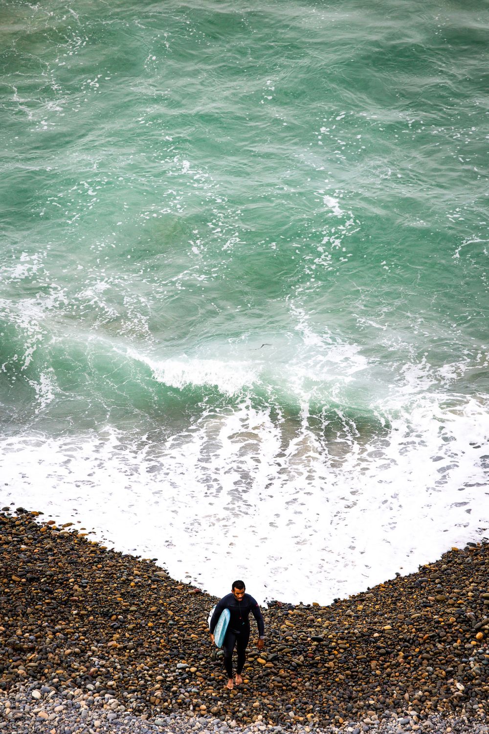 A lone figure climbs up a rocky beach as turquoise waves crash against the shore.