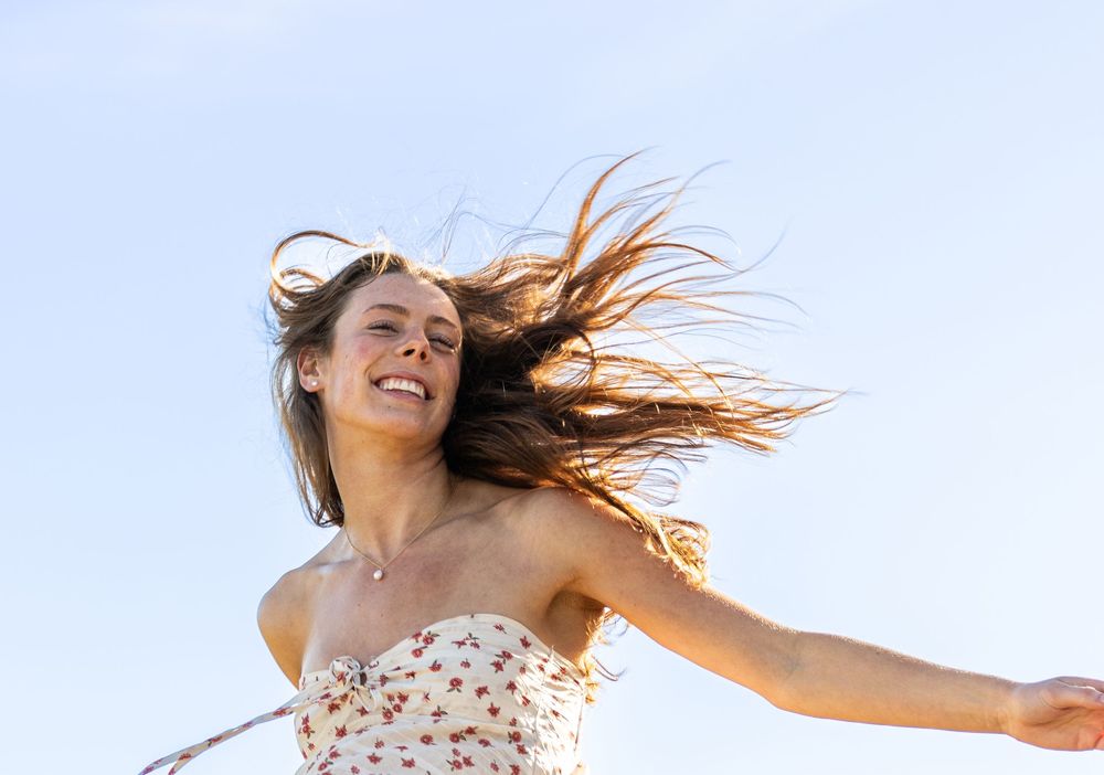 Joyful young woman in floral sundress with hair blowing in wind against blue sky, arms outstretched.