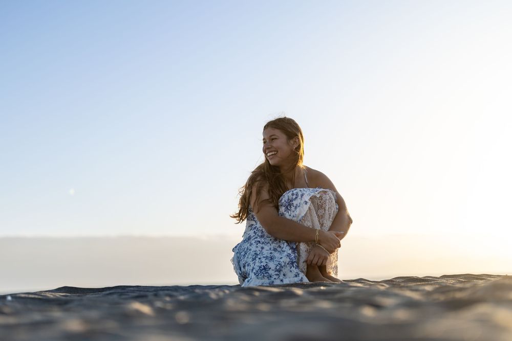 A joyful young woman in a floral dress sits on a sandy beach at sunset, laughing and enjoying the warm golden light.