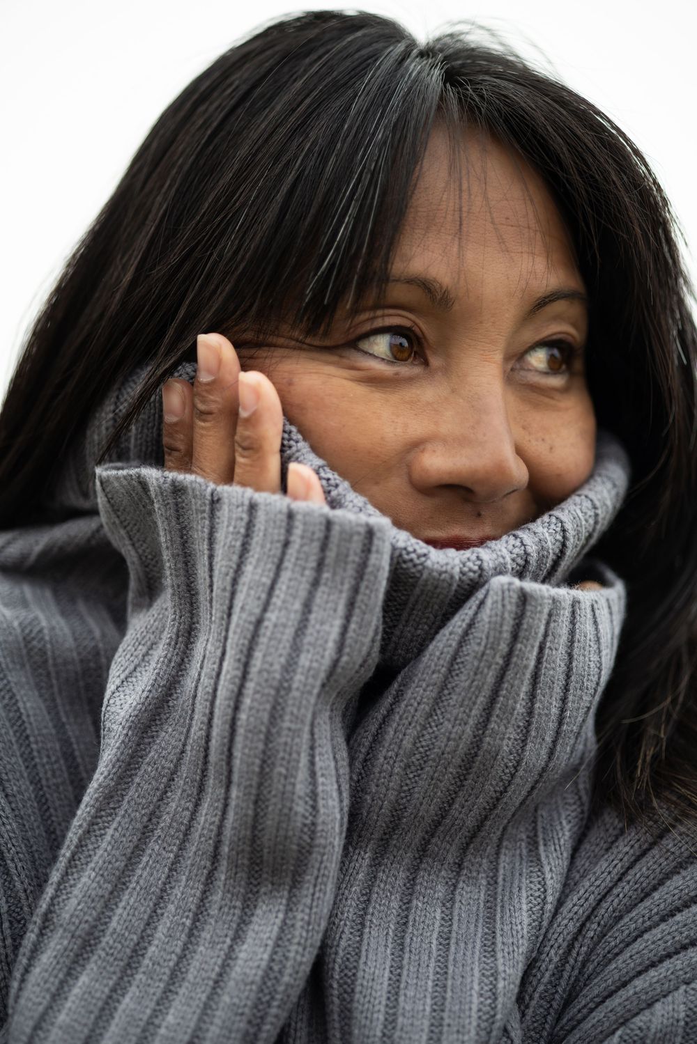 A middle-aged Asian woman in a gray turtleneck sweater pulls the collar up to her face while looking sideways.