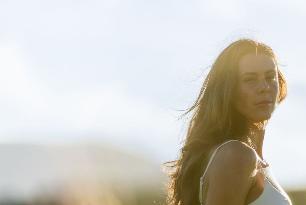 Young woman with long hair glowing in golden sunlight, looking over her shoulder outdoors.