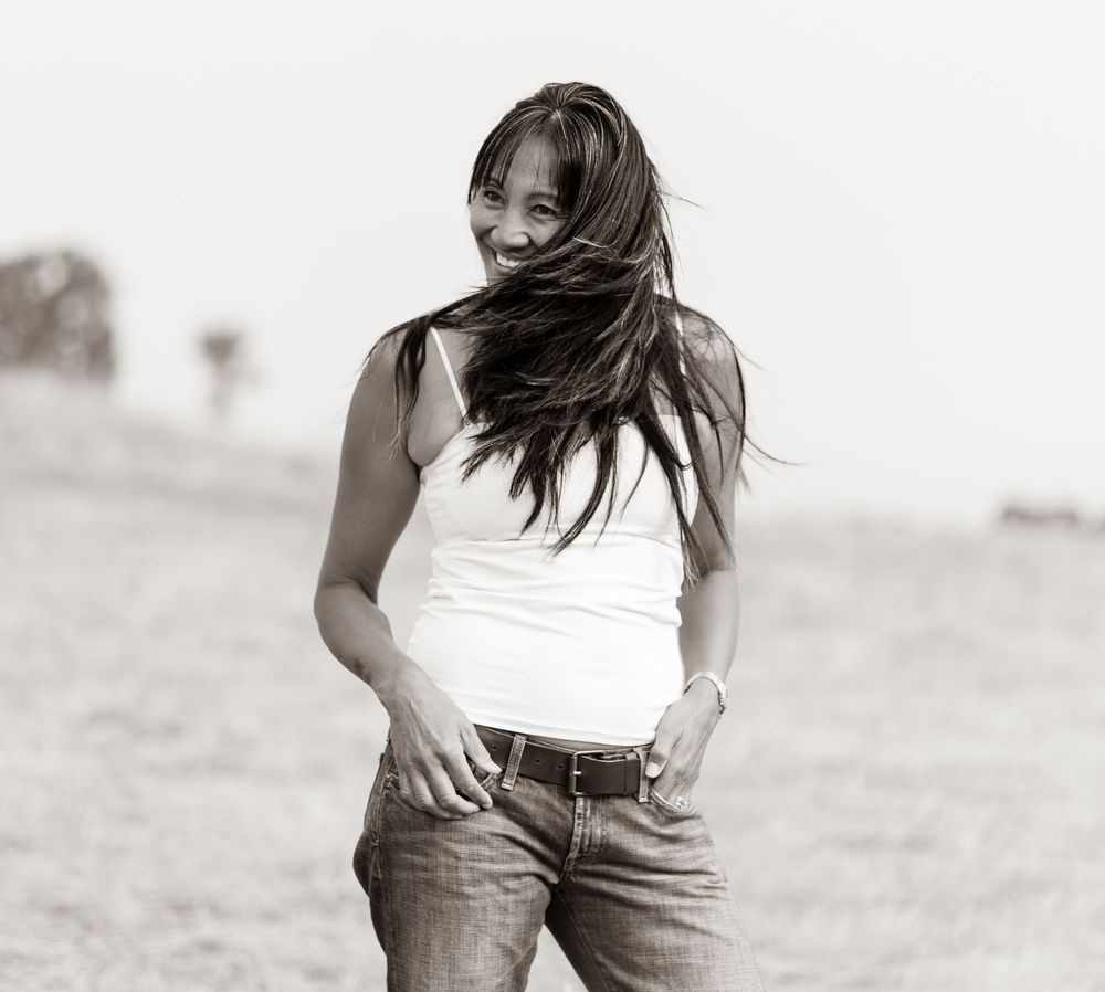Black and white photo of a smiling woman with windswept hair wearing a white tank top and jeans outdoors.