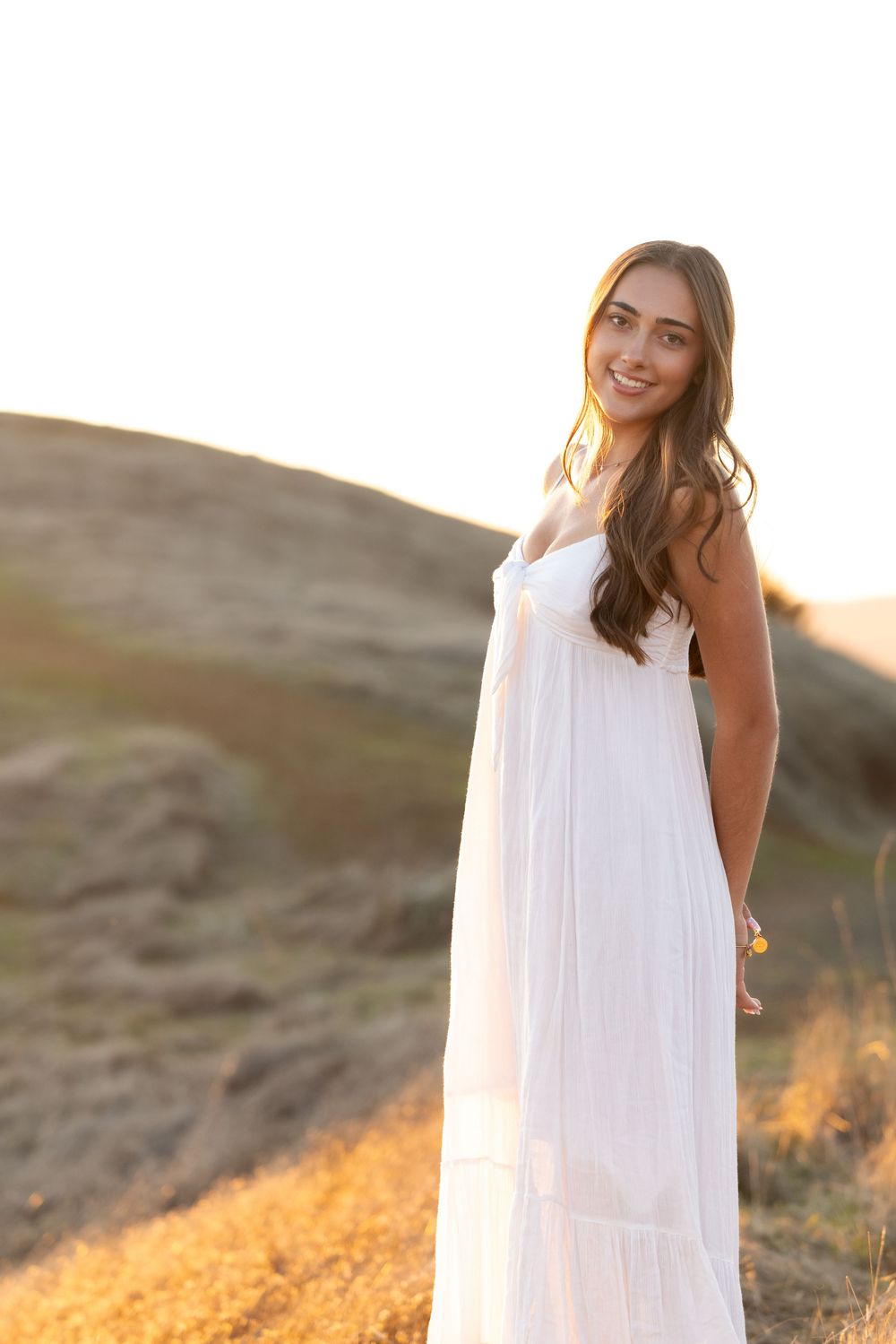 A person in a flowing white sundress poses on a hillside during golden hour sunset.