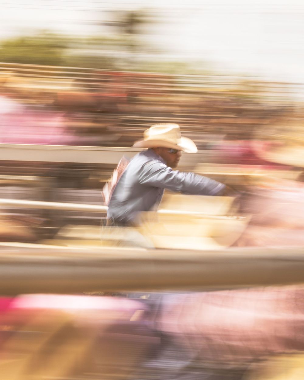 Blurred motion photo of a cowboy in a straw hat at a rodeo event with colorful crowd in background.