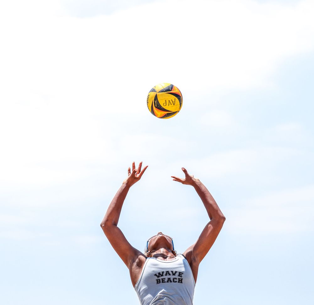 A volleyball player in a white jersey jumps high to set a yellow ball against a bright blue sky.