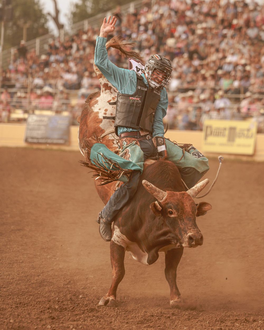 A bull rider in teal shirt waves one hand while riding a bucking bull at a packed rodeo arena.