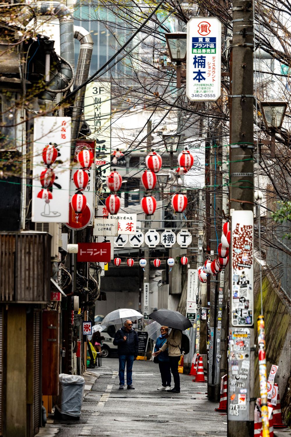 Narrow Japanese alley with red paper lanterns, signs, and people with umbrellas on a rainy day.