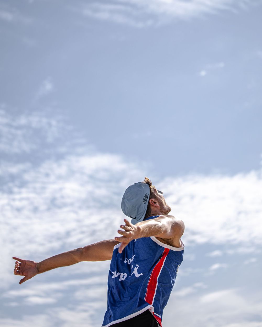 Male athlete in blue jersey throwing a frisbee against a partly cloudy sky outdoors.